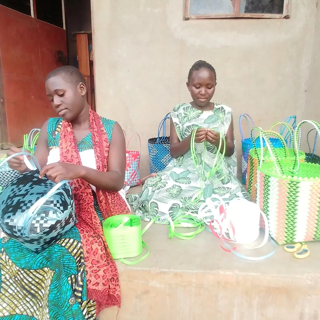 Girls seated outside weaving handles onto colorful handmade bags.