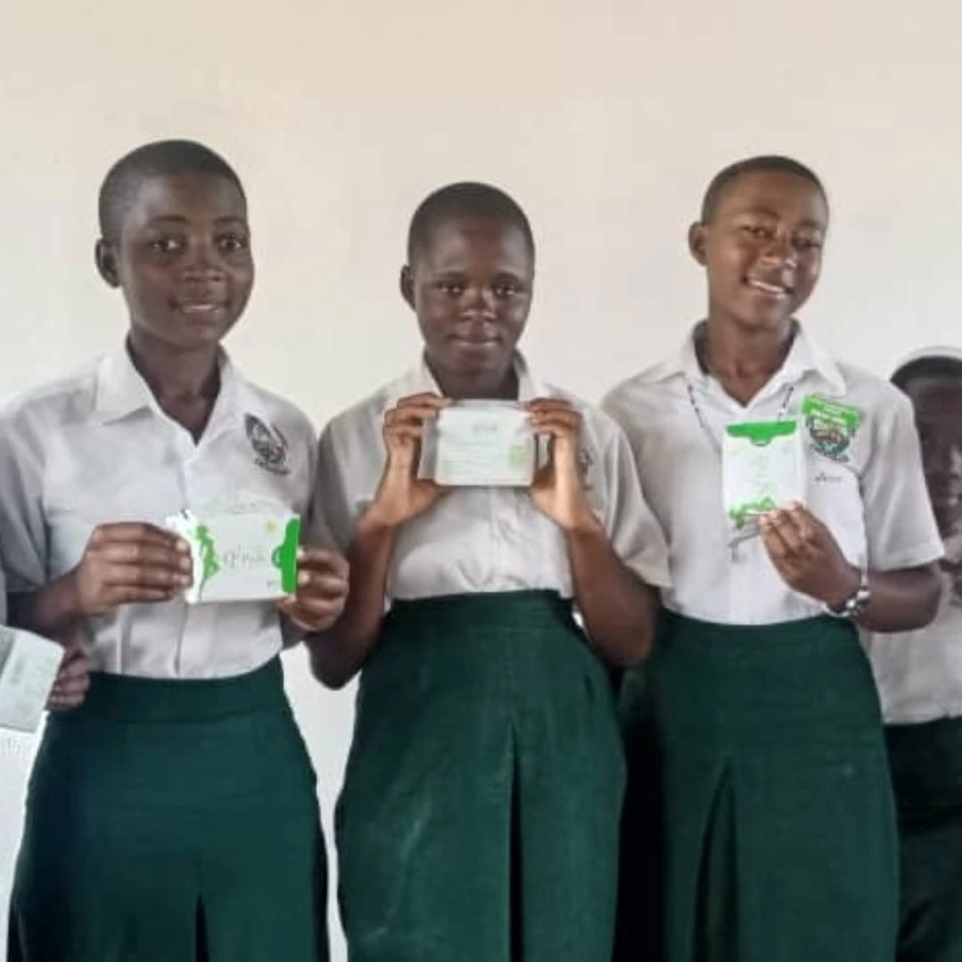 Three secondary school girls in green uniforms standing indoors and holding reusable menstrual pad packages during a school health initiative.
