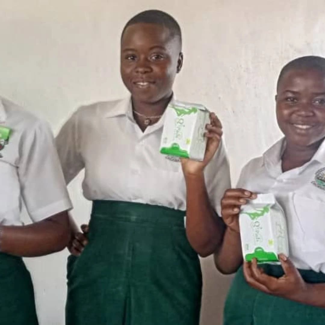 Two schoolgirls in uniform proudly holding reusable menstrual pad packages while standing against a plain classroom wall.