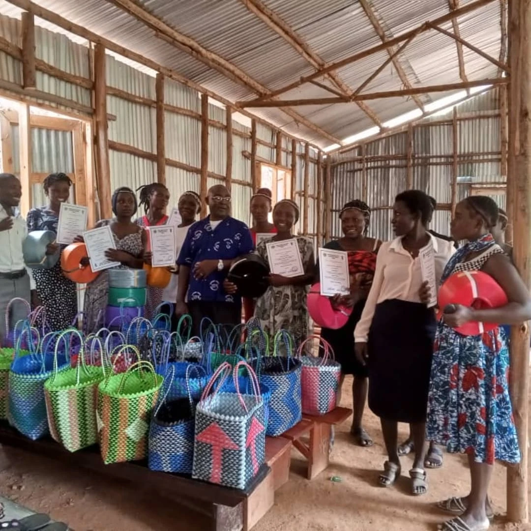 Women and girls holding certificates and handmade woven bags inside a community workshop.
